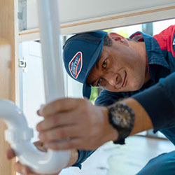 Goettl technician in branded navy cap with red band working on P-trap plumbing under sink.