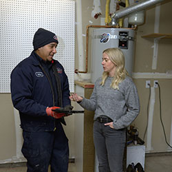 Goettl technician in navy uniform with Goettl logo holding tablet and consulting with female customer in front of a tank-style water heater in a residential garage.