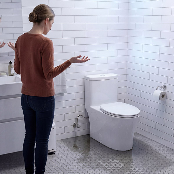 A woman standing in a bathroom with water pooled on the floor from a toilet leak, gesturing with frustration.